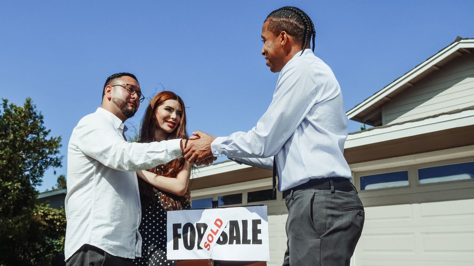 A real estate agent shaking hands with a happy buyer couple in front of their newly purchased home with a sold sign, celebrating the closed deal