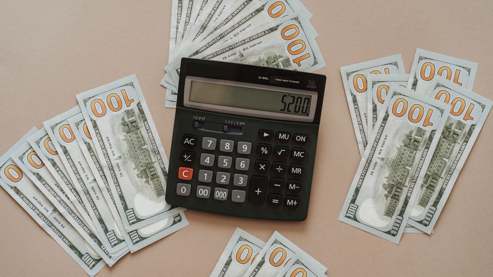 Top-down flat lay of a black desktop calculator reading 5200 surrounded by fanned U.S. one-hundred dollar bills on a warm tan background