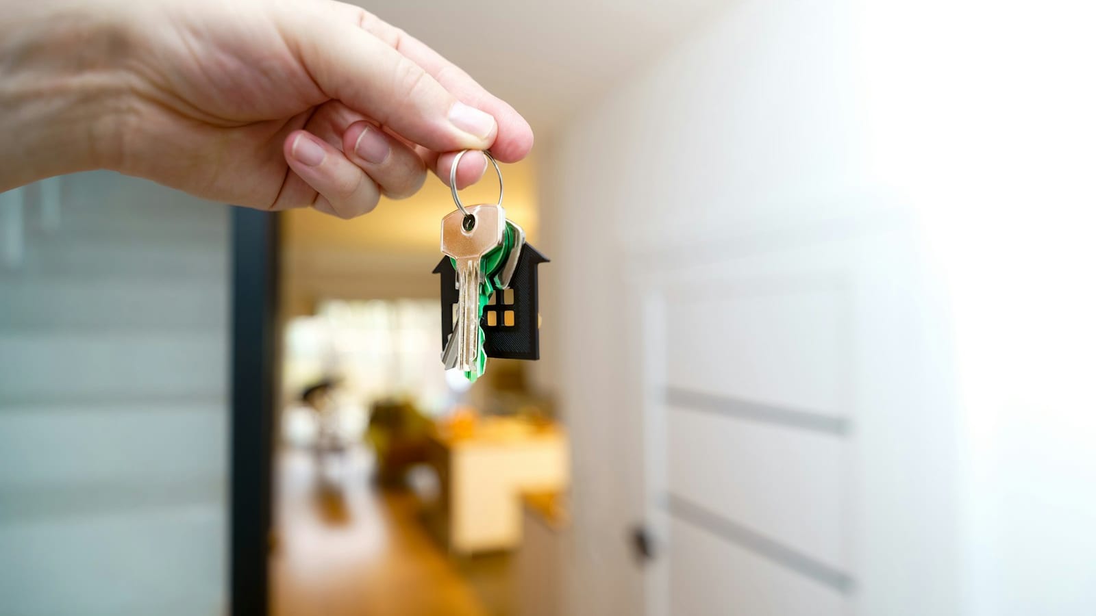 A hand holding house keys with a house-shaped keychain in front of a warmly lit home