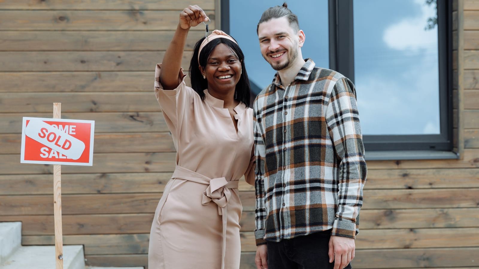 Happy young couple standing outside their new home, the woman joyfully holding up a house key next to a SOLD sign