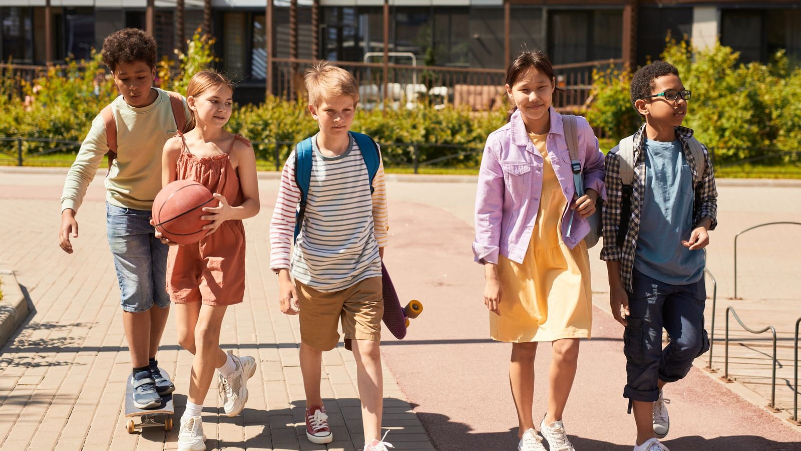 Five diverse elementary-age children walk to school together carrying backpacks, a basketball, and a skateboard on a sunny day in front of a modern residential building